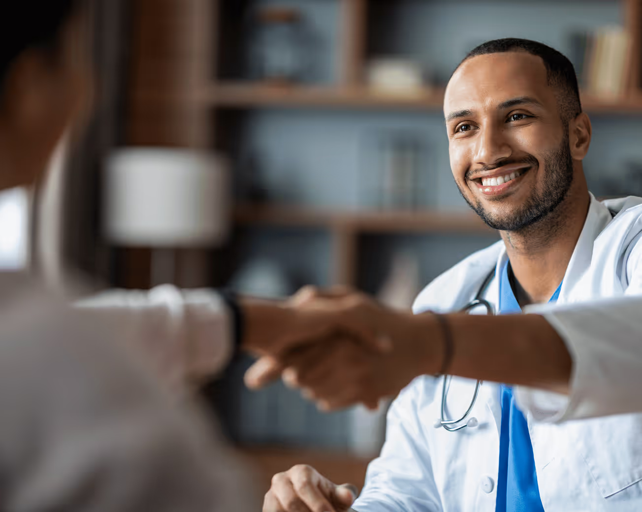 Smiling male doctor wearing a white coat and stethoscope shaking hands with a patient in an office.